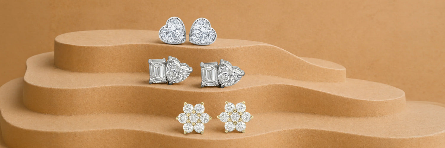 Lab-grown diamond stud earrings — heart cut halo studs in white gold, mixed emerald and heart cut two-stone studs in white gold, and round brilliant floral cluster studs in yellow gold on tiered camel sand display