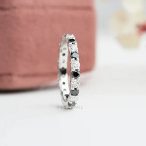 A close-up image of a Round Cut Lab Diamond Eternity Wedding Band featuring white and black lab-grown diamonds ring. It displayed against a soft pink background.