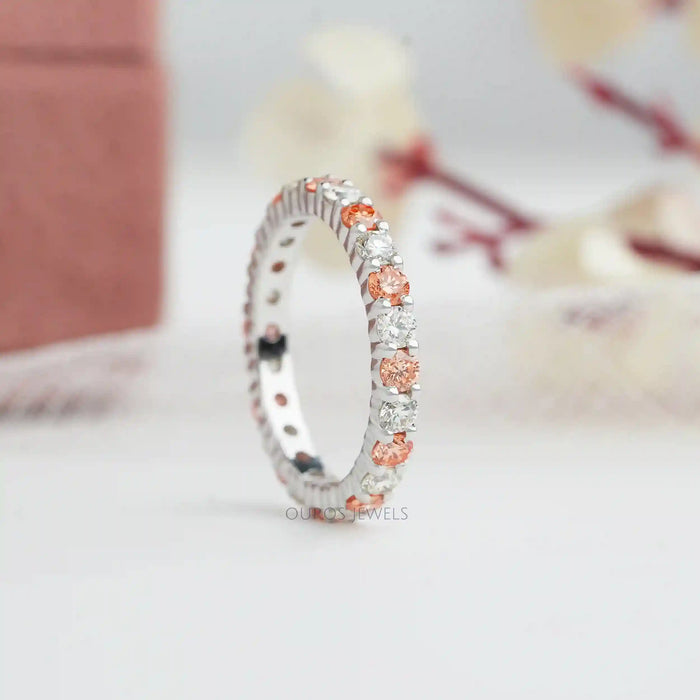 Round Cut Lab Diamond Eternity Wedding Band displayed upright on a white surface, featuring alternating white and pink lab-grown diamonds set around the entire band in a soft-focus background.